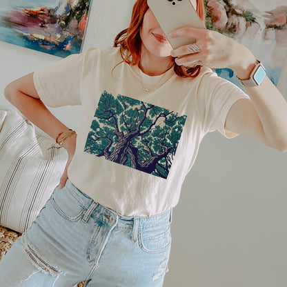 A woman takes a mirror selfie wearing a cream-colored t-shirt with a large green and blue tree graphic on the front, paired with light-wash distressed jeans.