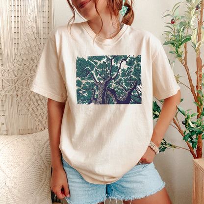 A woman wearing a cream-colored t-shirt with a square graphic print of a tree in green and dark blue, paired with light blue denim shorts in a cozy indoor setting.