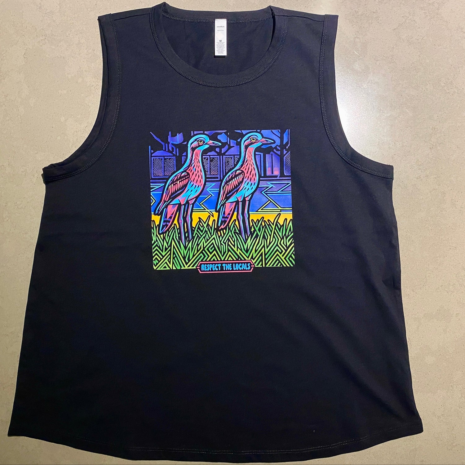 A black sleeveless tank top laid flat on a beige textured surface, featuring a colorful graphic print of two curlew birds in a stylized scene on the front.