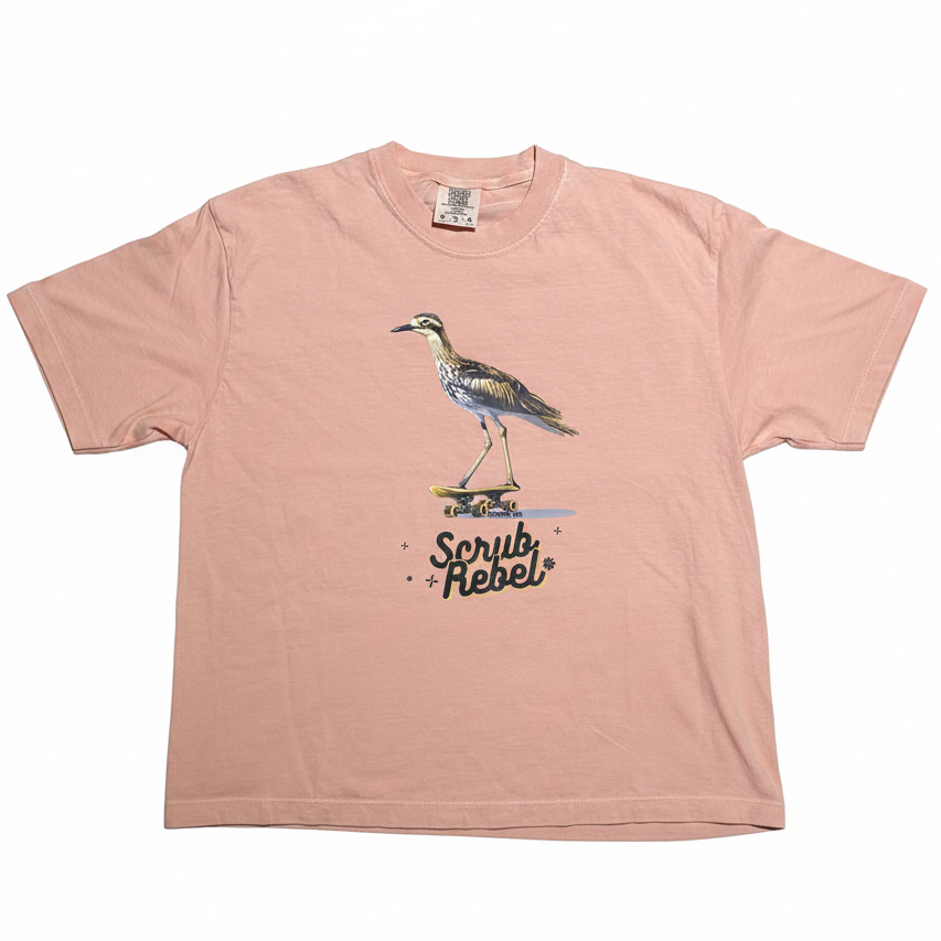 A light pink short-sleeve t-shirt with a graphic of a heron bird on a skateboard and the text 'Scrub Rebel' printed on the front, displayed flat against a white background.