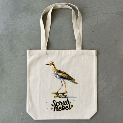 An off-white canvas tote bag with a printed illustration of a Curlew bird on a skateboard and the text 'Scrub Rebel' in black and gold, displayed flat on a gray concrete surface.