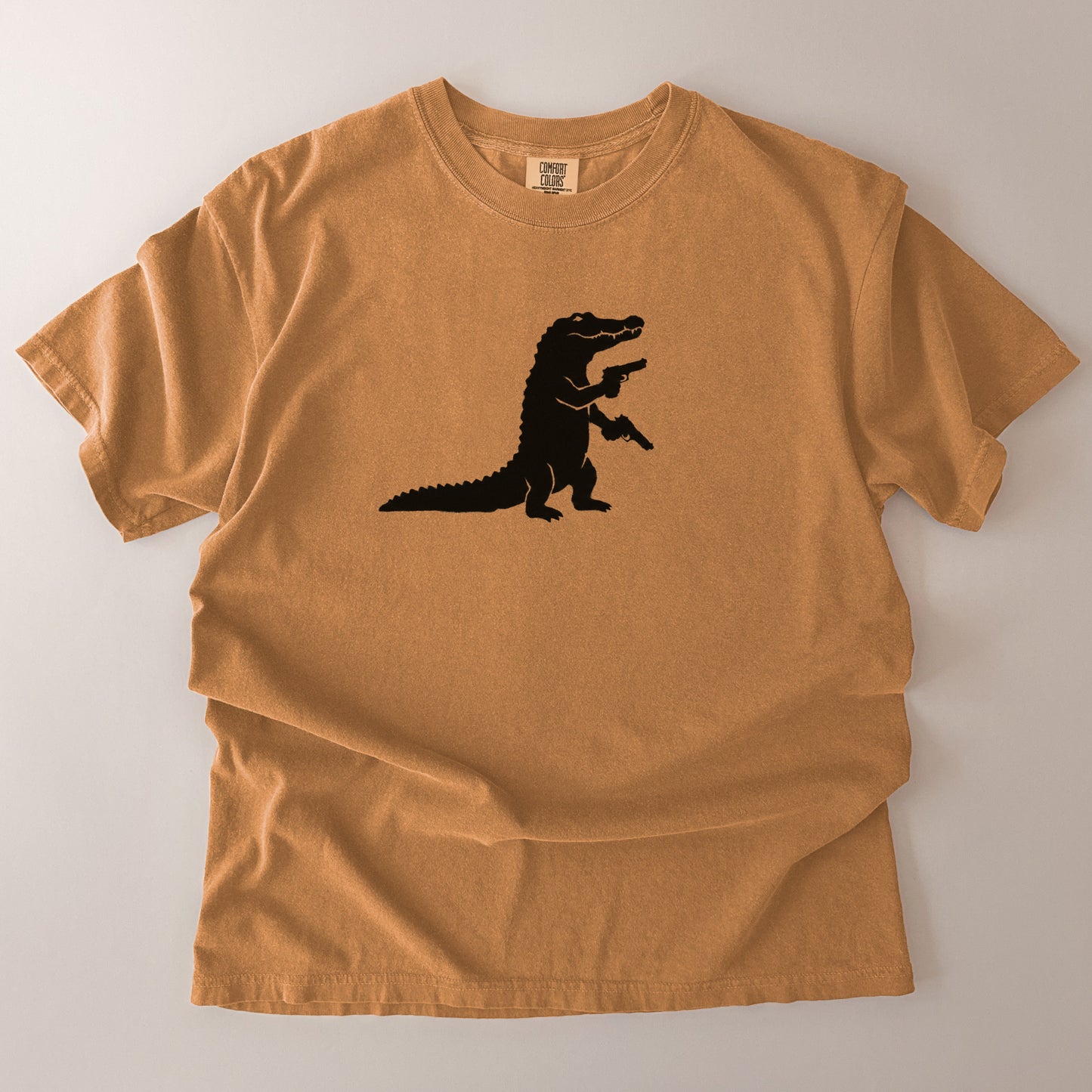 A tan crewneck t-shirt with a black silhouette of an alligator graphic on the chest, laid flat on a light gray background.