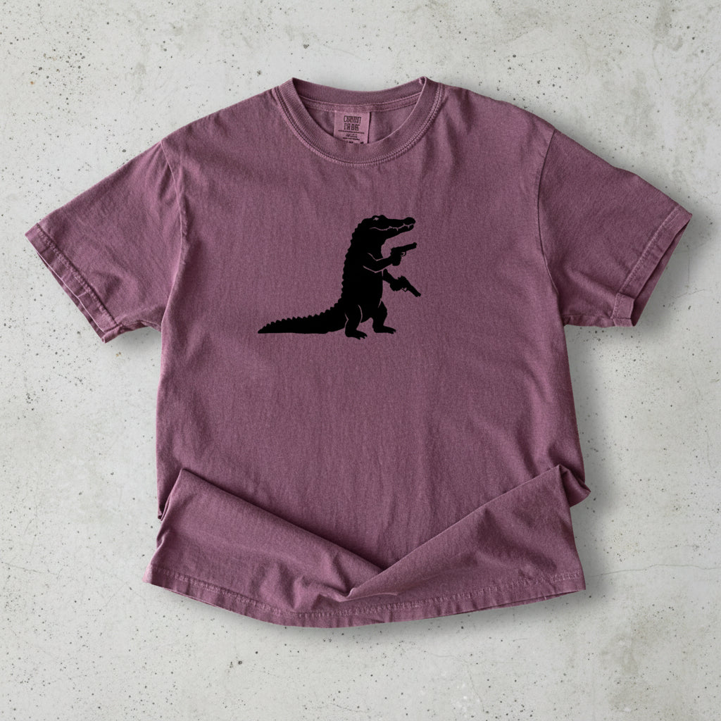 A short-sleeve crewneck t-shirt in a dusty mauve or plum purple color, featuring a black silhouette of a dinosaur-like creature on the chest. The shirt is laid flat against a white background with a soft drop shadow.