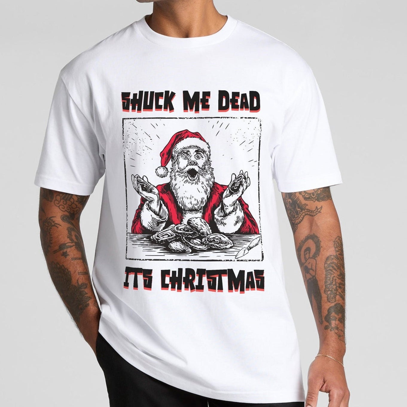 Person wearing a white t-shirt with a Christmas graphic design of santa shucking oysters saying shuck me dead its christmas text on a plain background