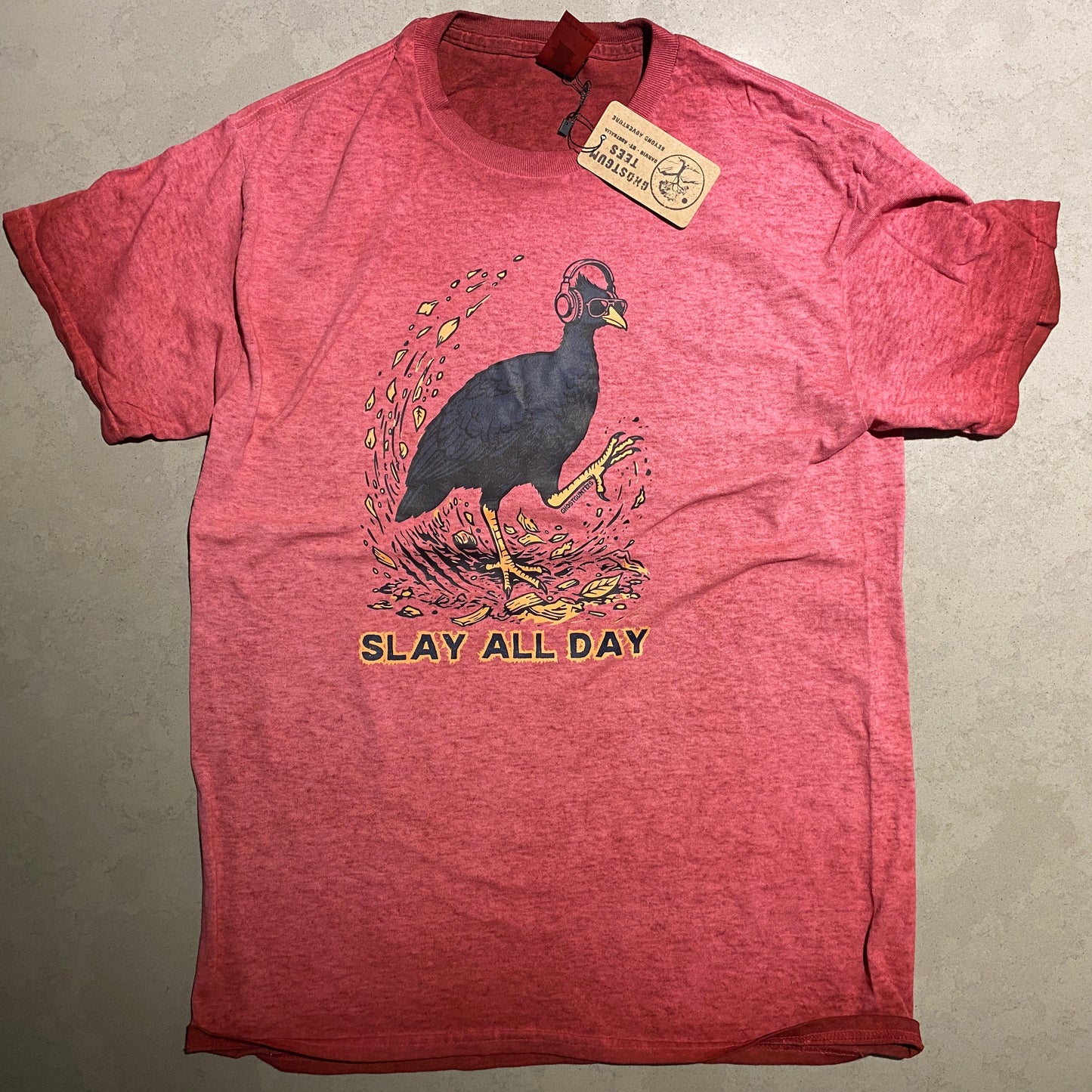 A heathered red short-sleeve graphic t-shirt laid flat on a gray surface. The shirt features a graphic of a bush chook with yellow accents and the text 'SLAY ALL DAY' printed on the front, with a paper brand tag still attached to the collar.