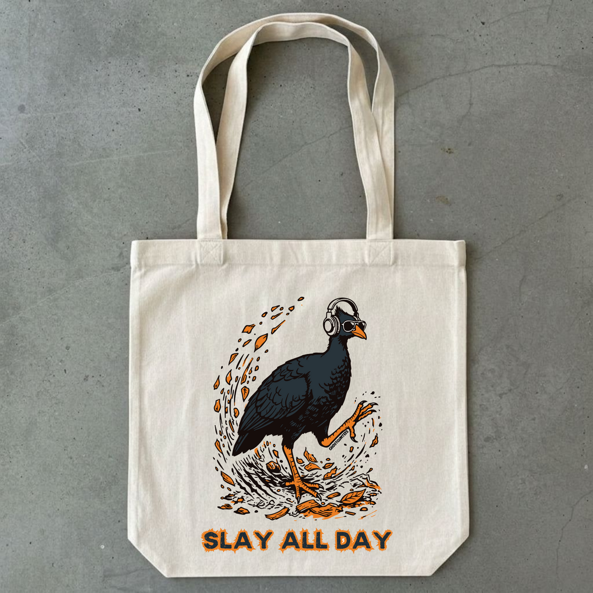 An off-white canvas tote bag with a graphic of a bush chook wearing headphones and orange text reading 'SLAY ALL DAY', photographed against a gray concrete background.