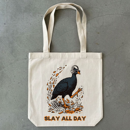 An off-white canvas tote bag with a graphic of a bush chook wearing headphones and orange text reading 'SLAY ALL DAY', photographed against a gray concrete background.