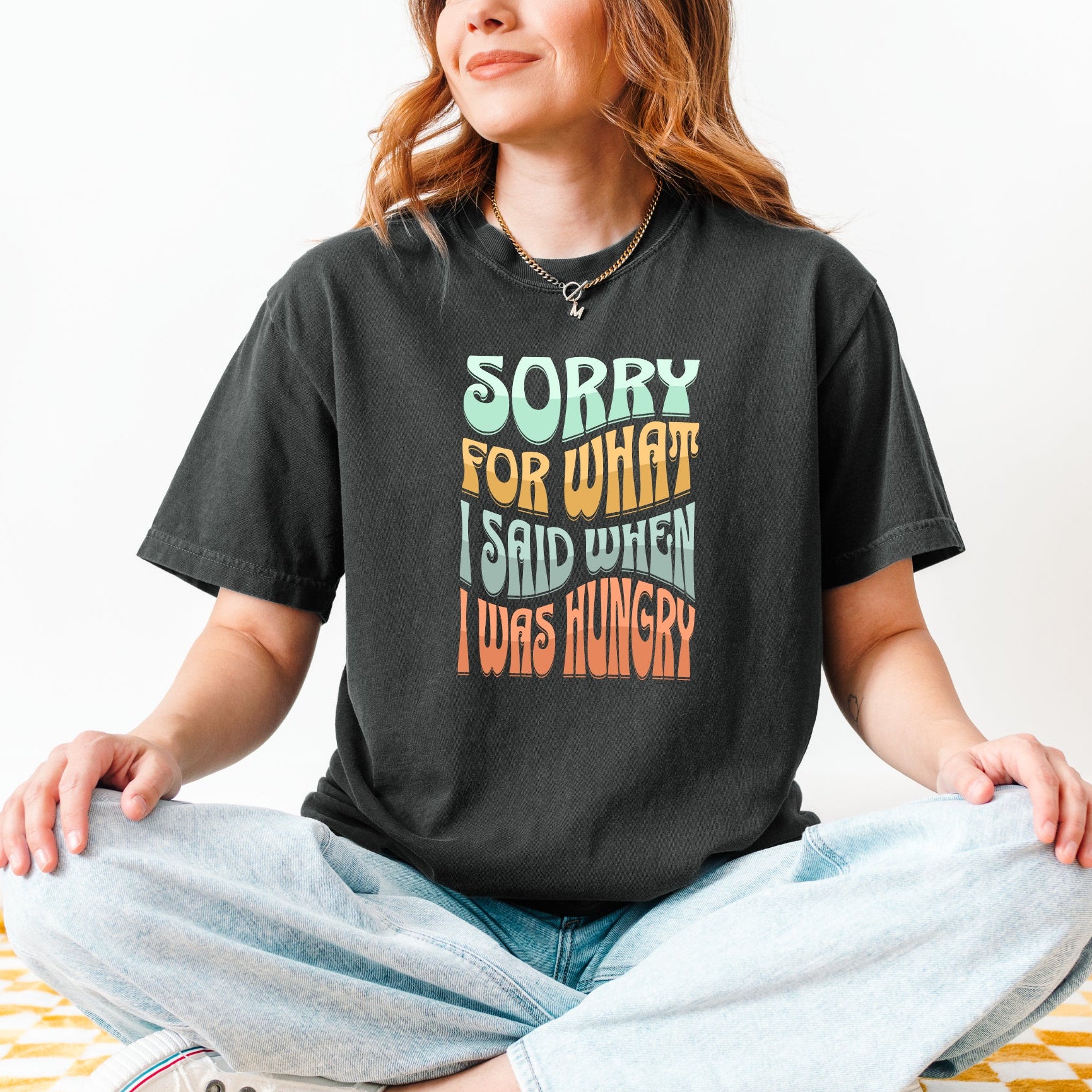 Person sitting cross-legged on a checkered floor wearing a black t-shirt with colorful text sorry for what i said when i was hungry.