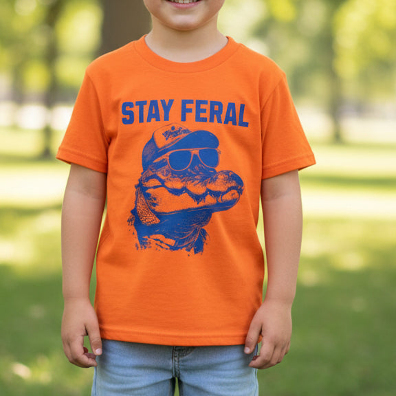 An orange short-sleeve t-shirt laid flat on a light wood floor, featuring a blue graphic of an alligator wearing sunglasses and a cap with the text 'STAY FERAL' printed above it.