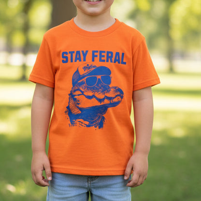 An orange short-sleeve t-shirt laid flat on a light wood floor, featuring a blue graphic of an alligator wearing sunglasses and a cap with the text 'STAY FERAL' printed above it.