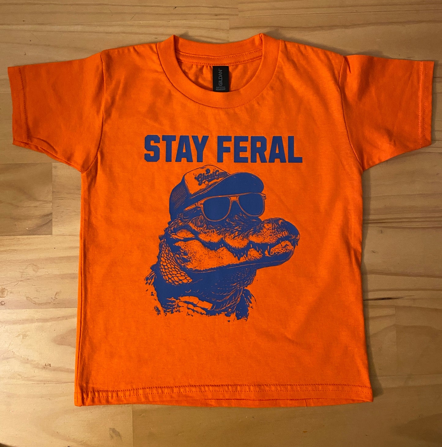 An orange short-sleeve t-shirt laid flat on a light wood floor, featuring a blue graphic of a turtle wearing sunglasses and the text 'STAY FERAL' printed vertically.