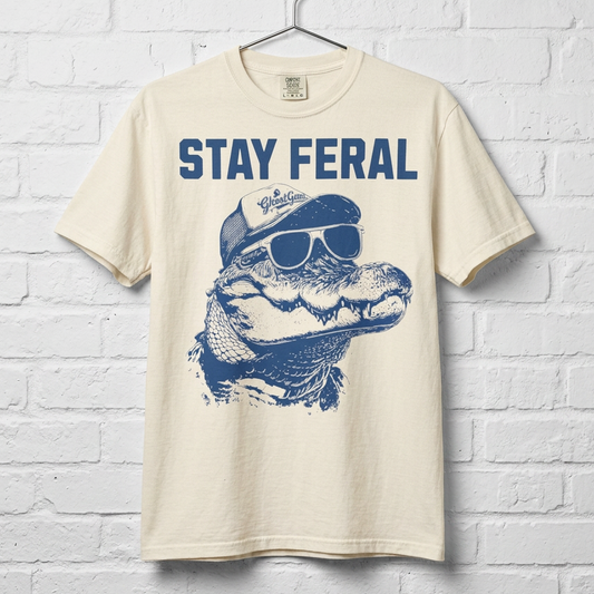 Beige t-shirt with blue graphic of an alligator and sunglasses, text 'Stay Feral' on a white brick wall background.
