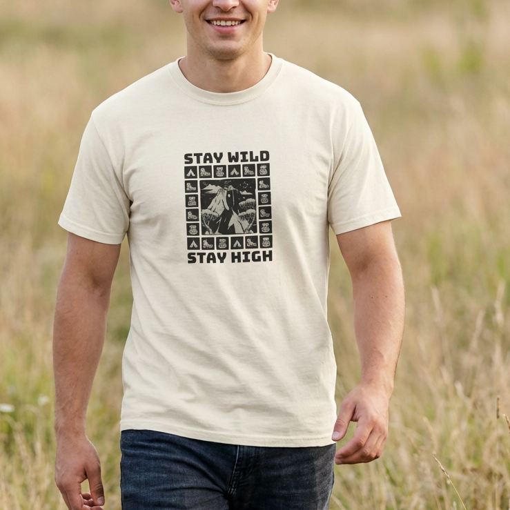 Man wearing a adventure t-shirt with 'Stay Wild Stay High' text and graphic in a grassy field