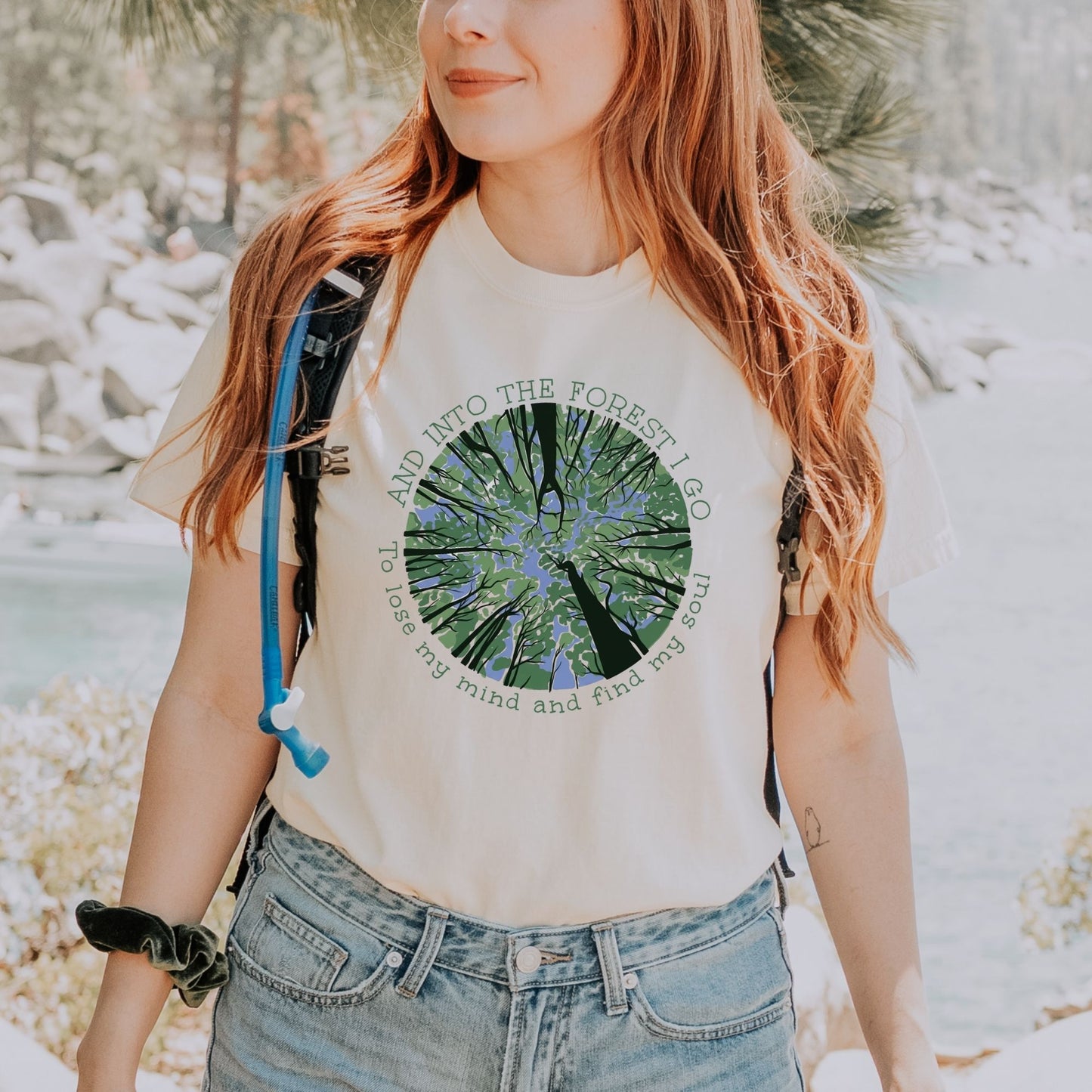 Woman wearing a white t-shirt with a nature forest graphic and text, standing in a forest.