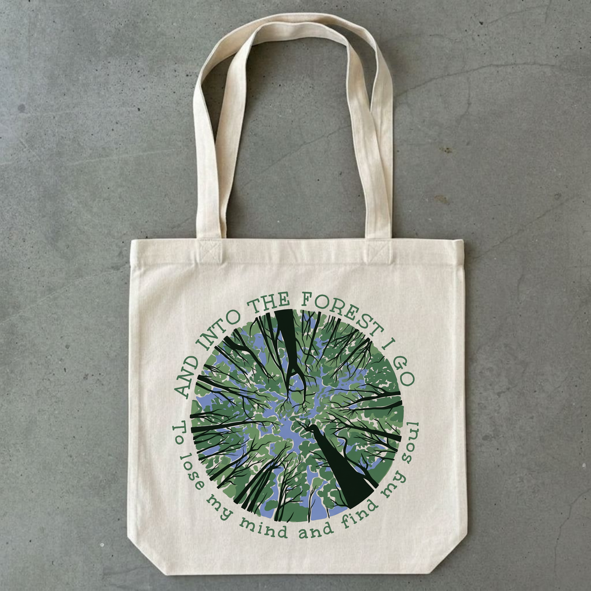 A natural canvas tote bag with a circular forest graphic in green, blue, and black, featuring the text 'AND INTO THE FOREST I GO To lose my mind and find my soul', displayed flat on a gray concrete surface.