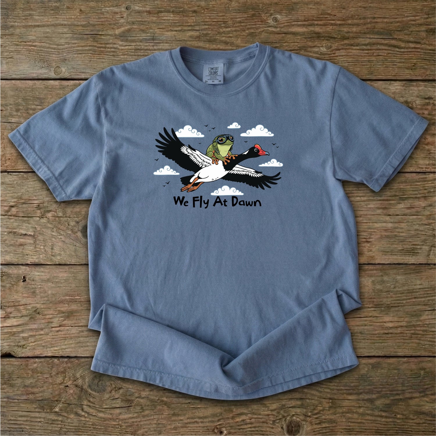 A dusty blue Ghost Gum Tees t-shirt featuring a humorous illustration of an Australian green tree frog wearing aviator goggles riding on the back of a flying magpie goose, with the text We Fly At Dawn below it.