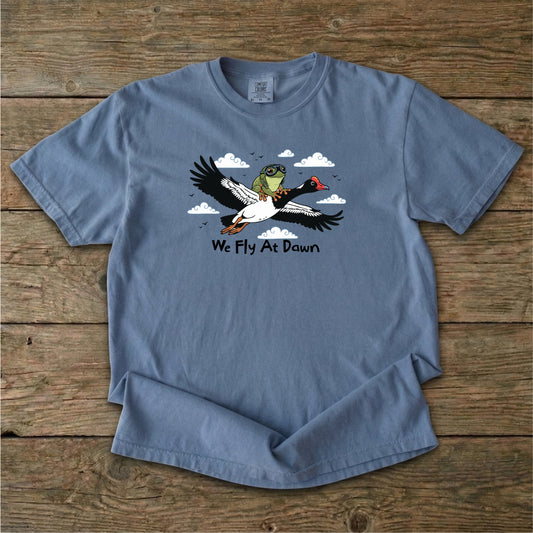 A dusty blue Ghost Gum Tees t-shirt featuring a humorous illustration of an Australian green tree frog wearing aviator goggles riding on the back of a flying magpie goose, with the text We Fly At Dawn below it.