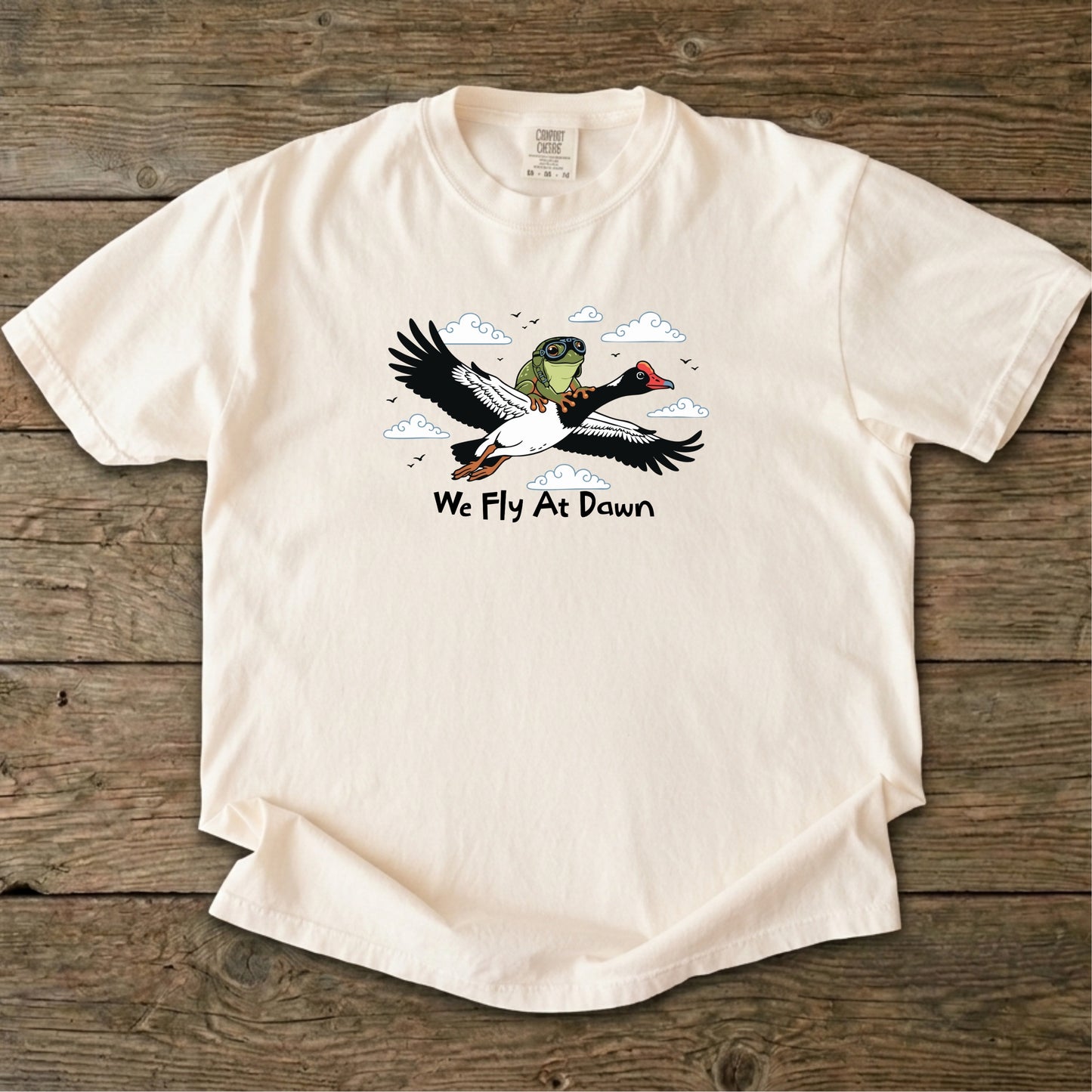 An Ivory Ghost Gum Tees t-shirt featuring a humorous illustration of an Australian green tree frog wearing aviator goggles riding on the back of a flying magpie goose, with the text We Fly At Dawn below it.