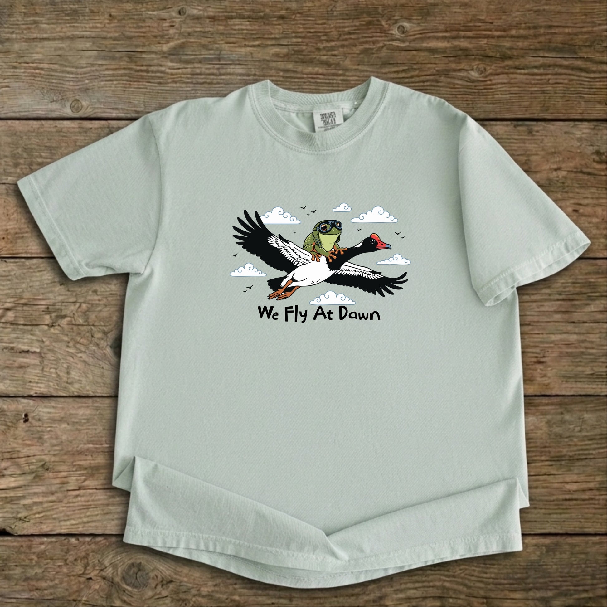 A dusty green Ghost Gum Tees t-shirt featuring a humorous illustration of an Australian green tree frog wearing aviator goggles riding on the back of a flying magpie goose, with the text We Fly At Dawn below it.