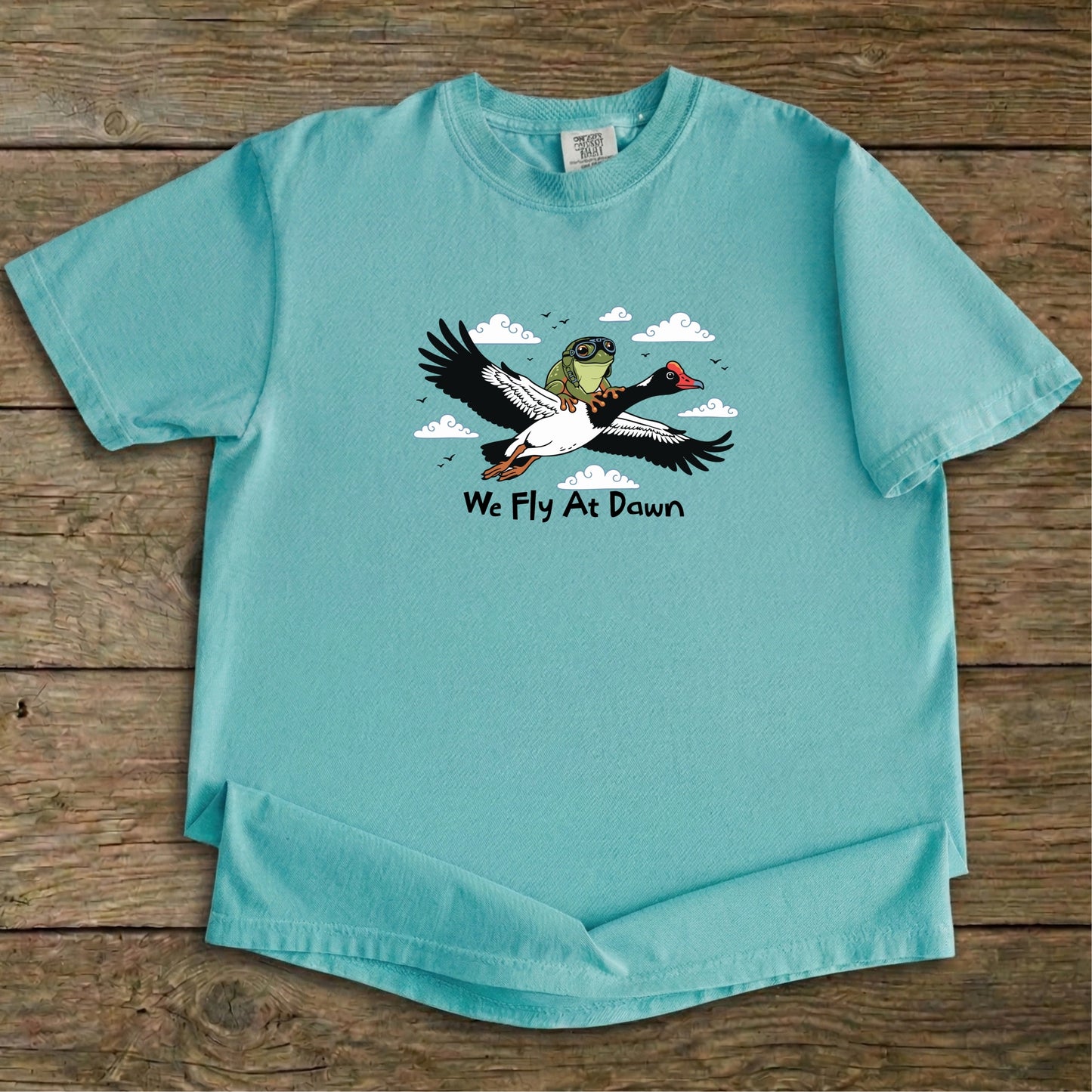 A light green Ghost Gum Tees t-shirt featuring a humorous illustration of an Australian green tree frog wearing aviator goggles riding on the back of a flying magpie goose, with the text We Fly At Dawn below it.