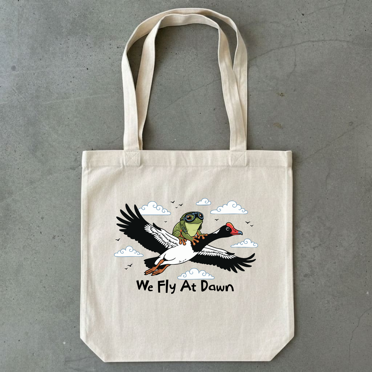 A natural canvas tote bag with a printed graphic of a goose and a green tree frog flying, with the text 'We Fly At Dawn' underneath, displayed flat on a gray concrete surface.