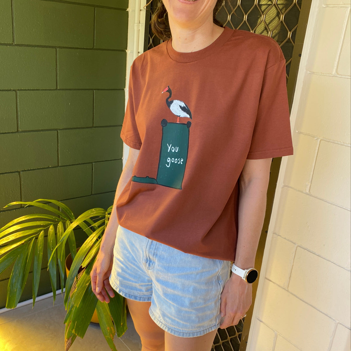 A woman wearing a rust-colored graphic t-shirt with a green and white bird design that says 'You goose', paired with light denim shorts, standing in front of a green brick wall and white door.