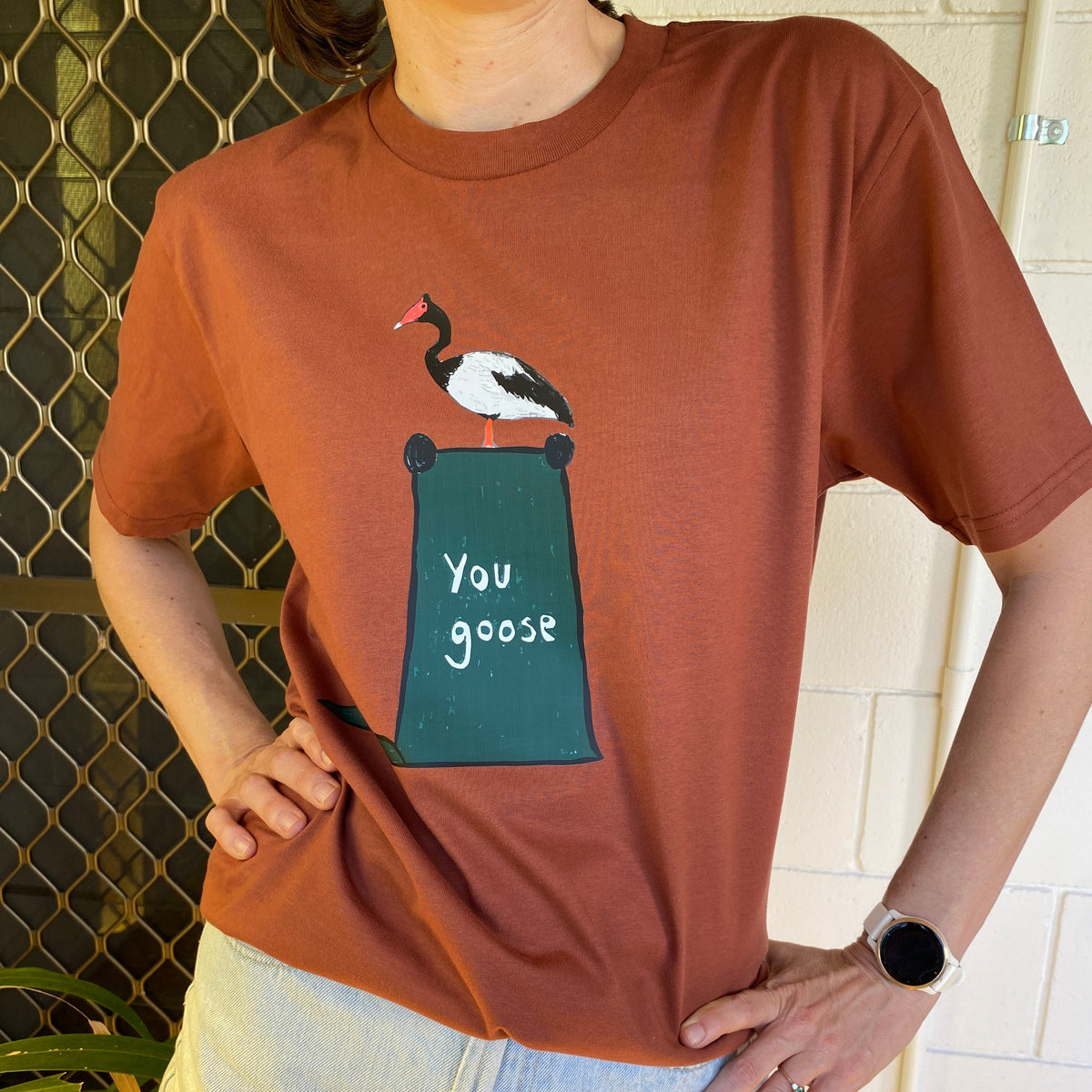 A person wearing a rust-orange graphic t-shirt with a goose design and the text 'You goose', paired with light-wash denim shorts, standing in front of a white brick wall.
