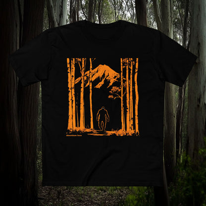 Silhouette of Bigfoot walking through tall trees with a mountain in the background, orange print on black T-shirt, outdoor adventure design by GhostGum Tees