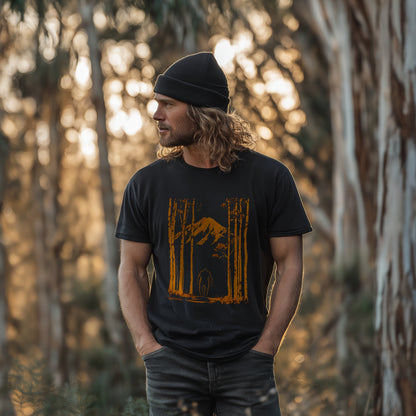 Bigfoot T-shirt with orange Yowie forest and mountain design, worn by a man with long hair and a beanie standing in the Australian bush at golden hour – GhostGum Tees.