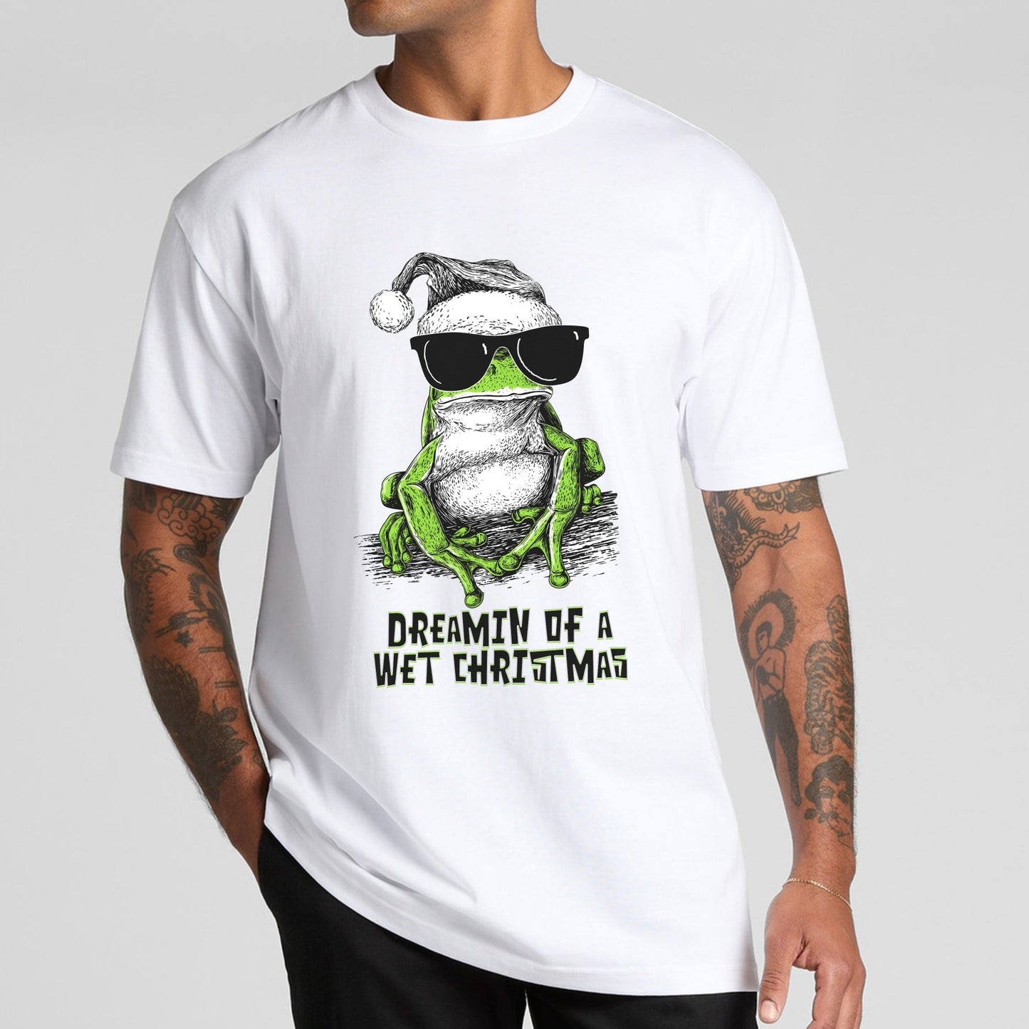 Green tree frog Christmas T-shirt — illustrated frog wearing sunnies and a Santa hat with the text “Dreamin’ of a Wet Christmas” printed on a white tee.