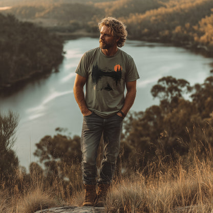 Man wearing a sage green kayaking T-shirt with lake and mountain print, standing on a grassy hill overlooking a scenic lake at sunset -GhostGum Tees