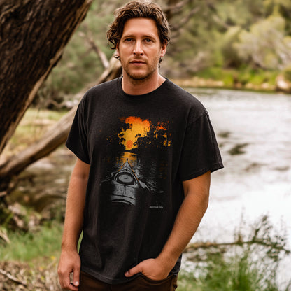 Man standing outdoors by a river, wearing a funky epic black T-shirt with a sunset kayaking scene printed on the front.