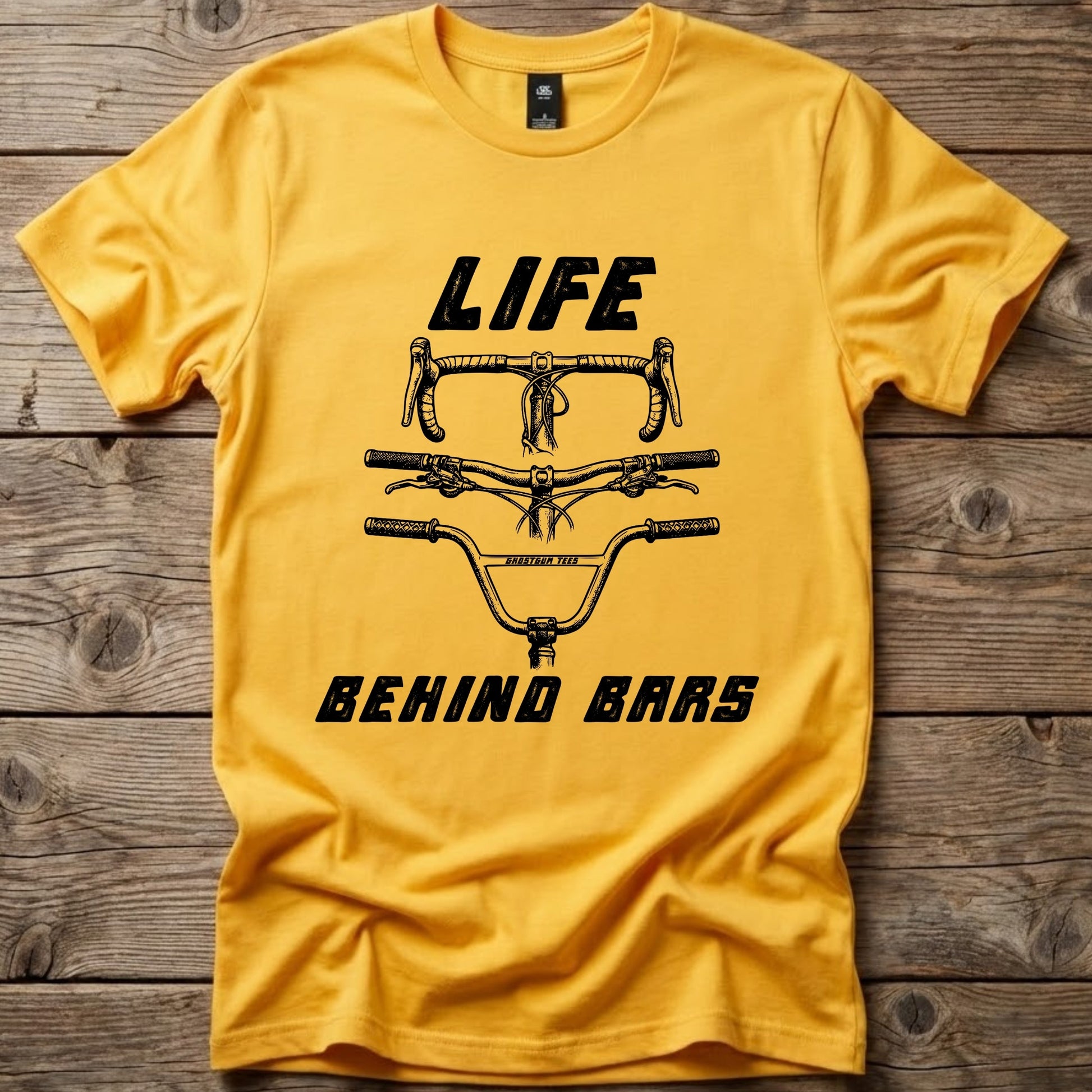 Yellow Life Behind Bars graphic t-shirt by Ghost Gum Tees folded on a rustic wooden background, featuring a black illustration of drop, mountain bike, and BMX handlebars.