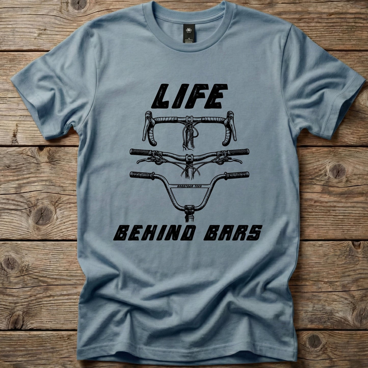 Blue Life Behind Bars graphic t-shirt by Ghost Gum Tees folded on a rustic wooden background, featuring a black illustration of drop, mountain bike, and BMX handlebars.