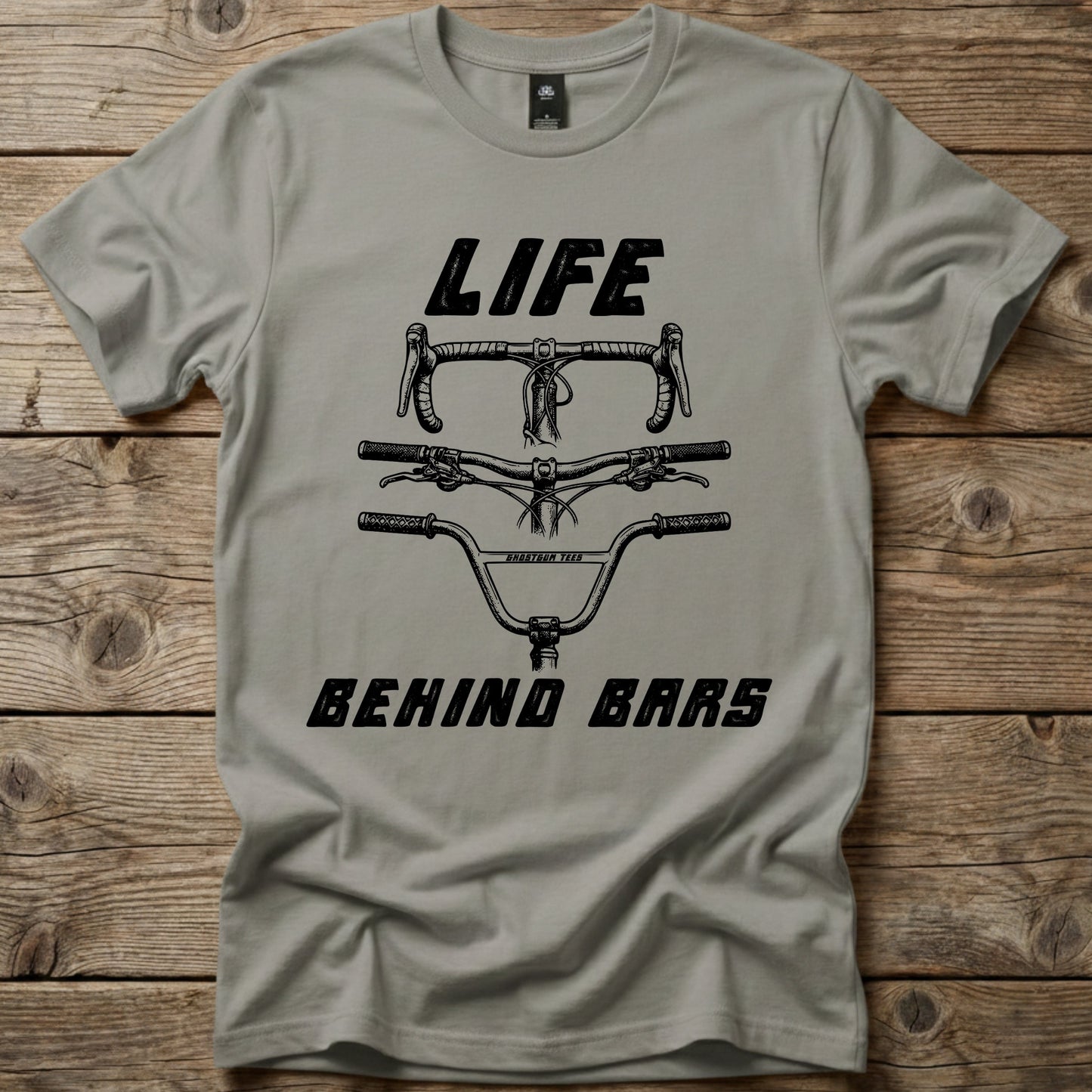 Grey Life Behind Bars graphic t-shirt by Ghost Gum Tees folded on a rustic wooden background, featuring a black illustration of drop, mountain bike, and BMX handlebars.