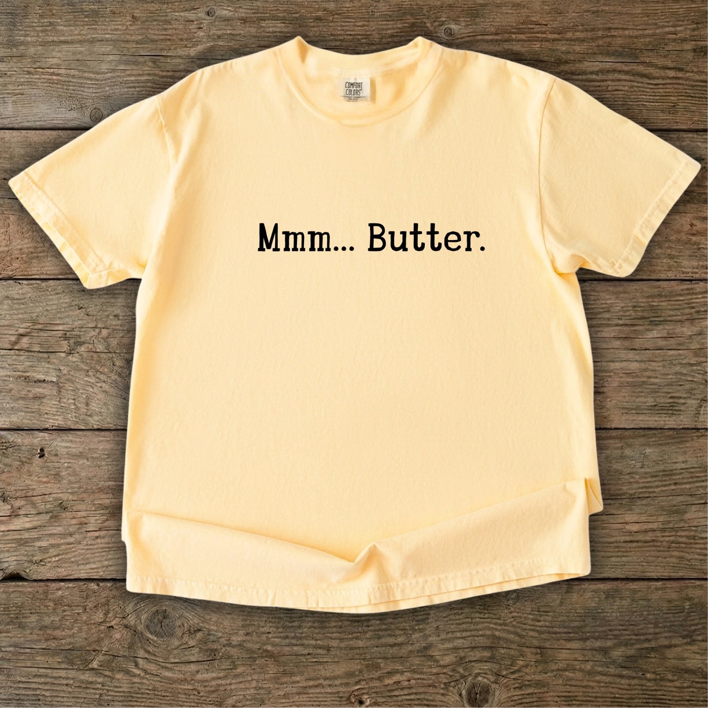Butter yellow vintage wash t-shirt featuring the text Mmm Butter in black, laid flat on a rustic wooden timber background.