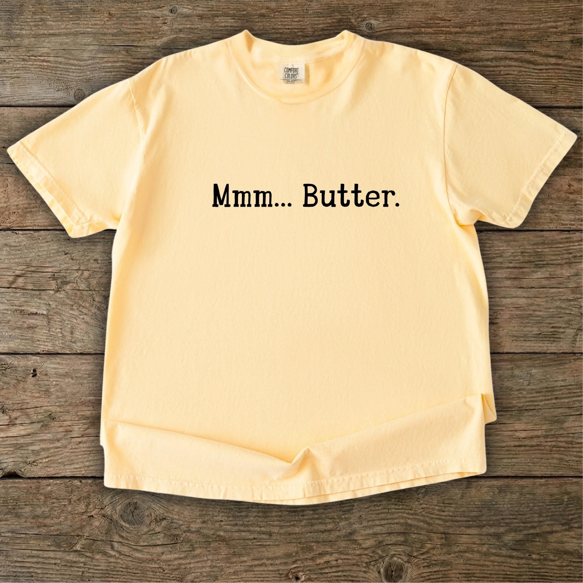 Butter yellow vintage wash t-shirt featuring the text Mmm Butter in black, laid flat on a rustic wooden timber background.