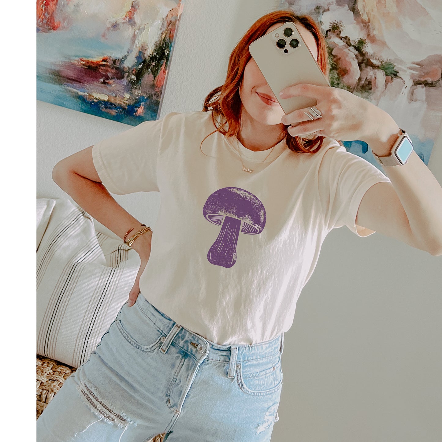 A woman takes a mirror selfie wearing a cream-colored t-shirt with a purple mushroom graphic on the front, paired with light-wash distressed jeans.
