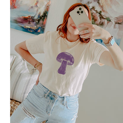 A woman takes a mirror selfie wearing a cream-colored t-shirt with a purple mushroom graphic on the front, paired with light-wash distressed jeans.