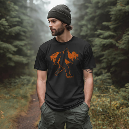 Man wearing a black GhostGum Tees "Take a Hike" t-shirt featuring an orange Bigfoot graphic walking across mountains, standing on a forest trail