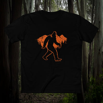 Black t-shirt featuring an orange Bigfoot silhouette walking across mountain peaks, displayed against a dense forest background